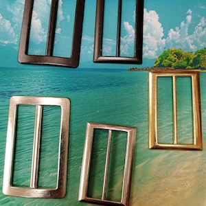 May include: An assortment of rectangular metal buckles in various finishes. The buckles include black, silver, and gold tones. Each buckle has a central bar and is designed for use in belts, bags, or other accessories. The buckles are displayed against a backdrop of a beach scene.
