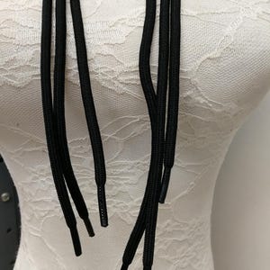 Könnte beinhalten: Nahaufnahme von mehreren schwarzen Schnürsenkeln mit Kunststoff-Aglets. Die Schnürsenkel sind dick und scheinen aus einem gewebten Material zu bestehen. Die Aglets sind glatt und zylindrisch. Der Hintergrund ist ein weißes, strukturiertes Gewebe.