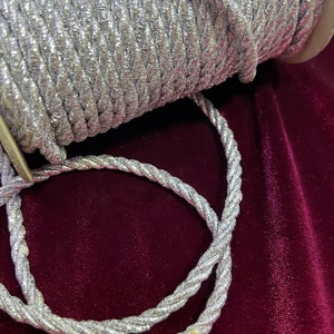 Puede incluir: Un carrete de cordón metálico plateado, con un primer plano del cordón en sí, mostrando su textura trenzada.