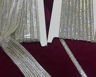 May include: A close-up of two rolls of silver rhinestone trim. The trim is made of small, square rhinestones that are sewn together in rows. The trim is on a white background.