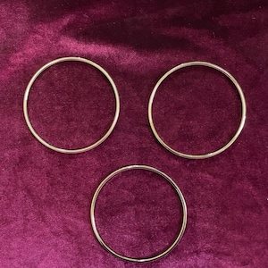 以下が含まれることがあります： 濃い赤紫色のベルベット生地の上に、3つの異なるサイズの銀色の金属製の輪が置かれています