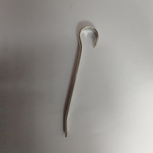 Puede incluir: Una herramienta de metal plateado y doblado, con un gancho curvado en la parte superior y un mango largo y delgado. La herramienta está diseñada con una ligera curva a lo largo de su longitud.