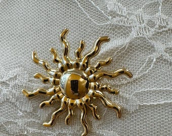May include: A gold-colored sunburst brooch with a central, reflective, circular element. The brooch features wavy rays extending outwards. The background is a white, textured fabric.