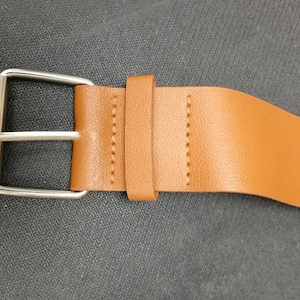 May include: A tan leather belt with a silver buckle. The belt features a rectangular buckle and a matching leather loop to secure the end. Stitching is visible along the belt's edge.
