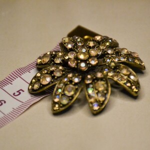 Puede incluir: Un broche dorado en forma de flor con pedrer&iacute;a iridiscente. El broche est&aacute; sobre una cinta m&eacute;trica rosa y blanca.