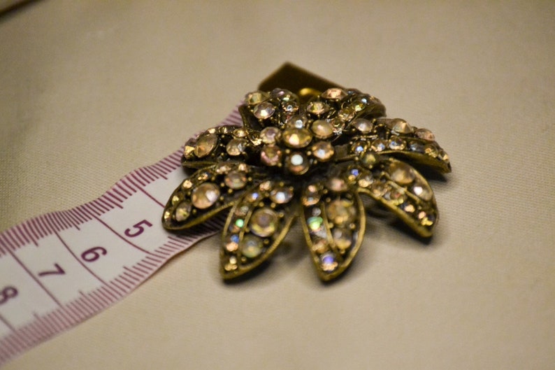 Puede incluir: Un broche dorado en forma de flor con pedrer&iacute;a iridiscente. El broche est&aacute; sobre una cinta m&eacute;trica rosa y blanca.