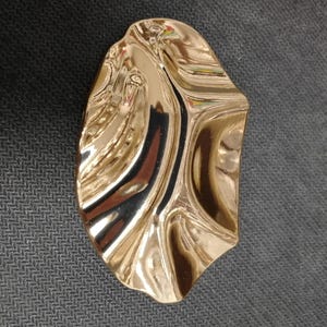 May include: A gold-coloured, abstract object with a rippled surface. The object has a glossy finish, reflecting light and creating highlights and shadows. The design is organic and fluid, suggesting a natural form.