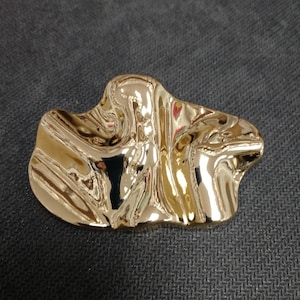 Può includere: Un oggetto metallico dorato di forma astratta con una superficie lucida e riflettente. Il design presenta curve organiche e fluide e una finitura liscia e lucida. L'oggetto è posto su uno sfondo scuro e strutturato.