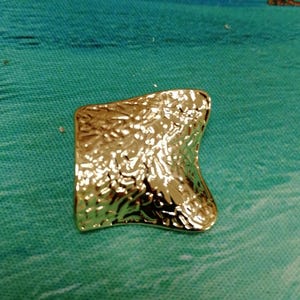 Peut inclure: Un objet doré de forme carrée et texturée, avec un bord légèrement incurvé. La surface présente un aspect martelé ou en relief, reflétant la lumière. L'objet est posé sur un fond bleu et turquoise.