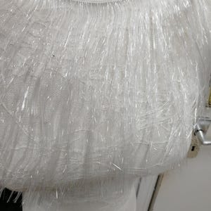 May include: Close-up of white fabric with a fringe of clear, thin, plastic-like strands. The fabric appears layered, with a lace-like texture visible beneath the fringe. The overall effect is delicate and textured.