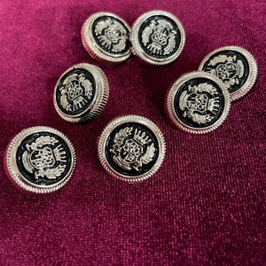 Puede incluir: Un conjunto de siete botones plateados y negros con un diseño decorativo. Los botones tienen un diseño en relieve de una corona y hojas.