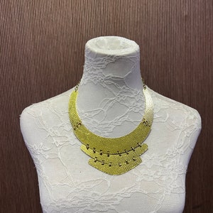 Op de afbeelding: Een gouden statement-ketting met een gelaagd ontwerp. De ketting heeft een breed, gebogen bovenstuk en drie kleinere, gelaagde stukken eronder. De ketting wordt getoond op een wit mannequin.
