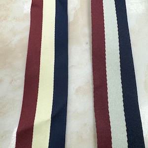 Op de afbeelding: Twee stoffen lintversieringen met drie strepen. Een lint is bordeauxrood, crèmekleurig en donkerblauw. De andere lint is bordeauxrood, crèmekleurig en zwart.