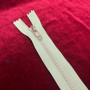 May include: A light green zipper with a silver slider. The zipper is lying on a red velvet surface.