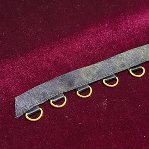 May include: A black fabric ribbon trim featuring a series of evenly spaced, small gold-toned metal D-rings attached with short fabric loops along one edge. The trim is presented against a deep burgundy velvet surface, highlighting the contrast.