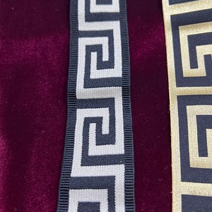 Puede incluir: Primer plano de dos cintas decorativas con un patr&oacute;n de greca. Una cinta es negra con dise&ntilde;o plateado, y la otra dorada con dise&ntilde;o negro. Sobre un fondo de terciopelo rojo oscuro.