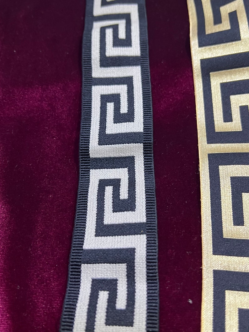 Puede incluir: Primer plano de dos cintas decorativas con un patr&oacute;n de greca. Una cinta es negra con dise&ntilde;o plateado, y la otra dorada con dise&ntilde;o negro. Sobre un fondo de terciopelo rojo oscuro.