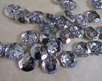 May include: A collection of silver-toned, faceted, two-hole buttons. The buttons are arranged in a random pattern on a light brown surface.