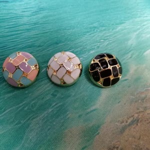 以下が含まれることがあります： 金色の縁取りが付いた、3つの丸い装飾ボタン。1つのボタンはパステルカラーブロックのデザイン、もう1つは白と金のパターン、3つ目は黒と金のパターンです。ボタンは、ターコイズとベージュの背景に配置されています。