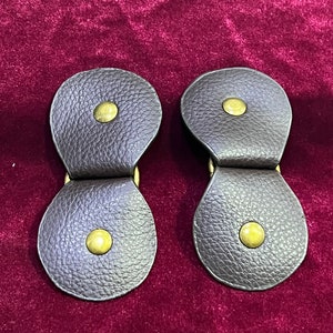 May include: Two brown leather toggles with brass buttons. The toggles are designed to be used as fasteners for clothing.