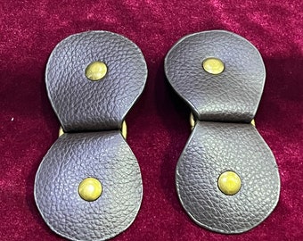 May include: Two brown leather toggles with brass buttons. The toggles are designed to be used as fasteners for clothing.