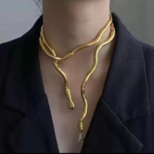 Op de afbeelding: Een goudkleurige slangenketting met een getextureerd, schubachtig ontwerp. De ketting wikkelt zich om de nek en drapeert over de voorkant, waardoor een stijlvolle en unieke look ontstaat. De kop en staart van de slang zijn zichtbaar.