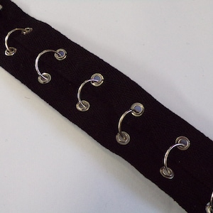Könnte beinhalten: Nahaufnahme eines schwarzen Stoffstreifens mit gleichmäßig verteilten, silberfarbenen Metallösen. Die Ösen sind gebogen und haben ein kleines, kreisförmiges Design. Der Stoff scheint ein robustes Material zu sein, möglicherweise für Kleidung oder Bastelprojekte.