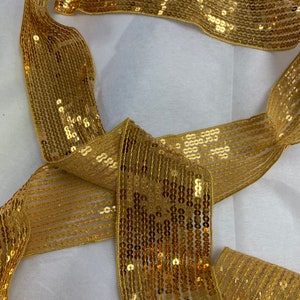 May include: Gold sequined ribbon with a sheer mesh backing. The sequins are arranged in rows and are a shiny gold color.