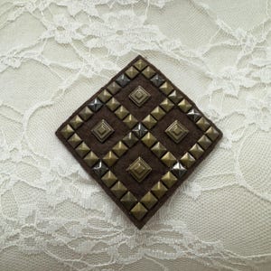 Op de afbeelding: Een bruine ruitvormige patch met een patroon van zilveren en gouden piramidevormige studs.