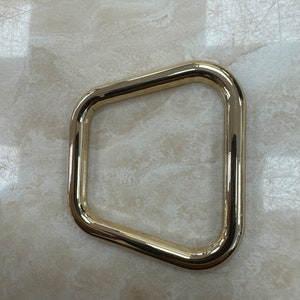 May include: A gold-colored, rectangular metal ring with rounded corners. The ring is a shiny, solid color and appears to be made of metal. The ring is a hardware component, likely for use in bags, belts, or other accessories.