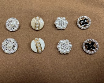 May include: Assortment of eight decorative buttons with pearl and rhinestone embellishments. The buttons are round and feature a variety of designs, including floral and geometric patterns. Some buttons have a black enamel center.
