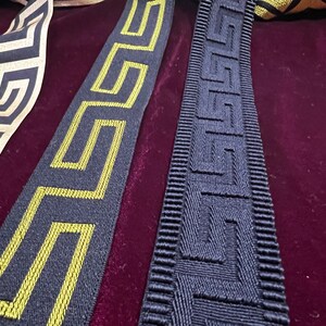 Puede incluir: Primer plano de cintas decorativas con patrones geom&eacute;tricos. Una cinta presenta un fondo negro con un dise&ntilde;o de llave griega dorada. Otra tiene un fondo negro con un dise&ntilde;o de llave griega amarilla. Una tercera cinta tiene un patr&oacute;n geom&eacute;trico blanco y dorado.