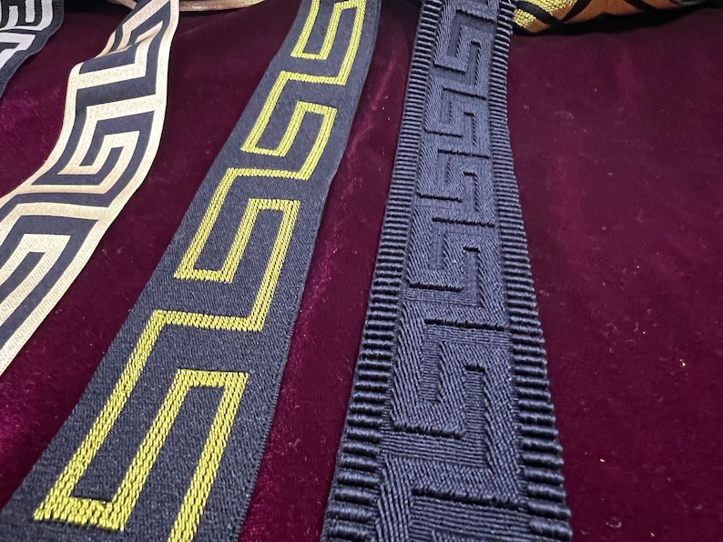 Puede incluir: Primer plano de cintas decorativas con patrones geom&eacute;tricos. Una cinta presenta un fondo negro con un dise&ntilde;o de llave griega dorada. Otra tiene un fondo negro con un dise&ntilde;o de llave griega amarilla. Una tercera cinta tiene un patr&oacute;n geom&eacute;trico blanco y dorado.