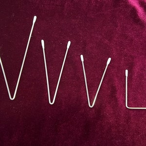 May include: Five silver metal bent wire shapes on a dark red velvet background. The shapes are all similar, but vary in size. The largest shape is a V, the next two are also Vs, but smaller. The next shape is a V, but even smaller. The smallest shape is a U.