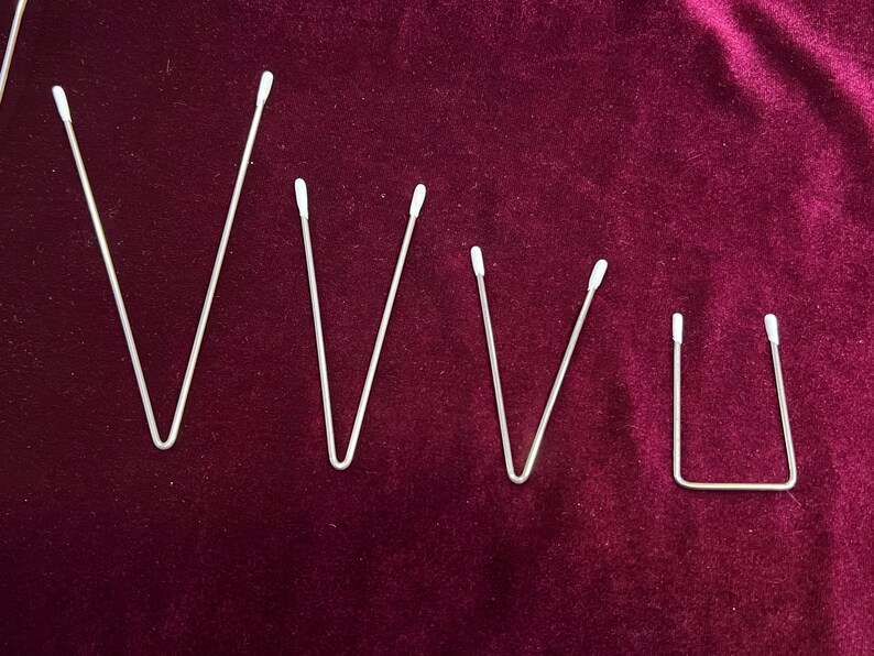 May include: Five silver metal bent wire shapes on a dark red velvet background. The shapes are all similar, but vary in size. The largest shape is a V, the next two are also Vs, but smaller. The next shape is a V, but even smaller. The smallest shape is a U.