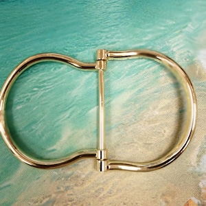 May include: A gold-colored metal belt buckle with a D-ring design. The buckle has a central bar connecting two rounded D-shaped loops. The background features a blurred image of a beach scene with turquoise water and sand.