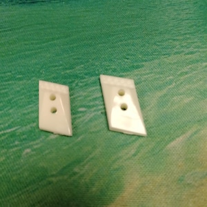 May include: Two white, asymmetrical, trapezoidal-shaped buttons. Each button features two vertical holes in the center and a textured, ridged pattern along the top slanted edge. The surface of the buttons is smooth and glossy with a solid white color.