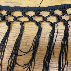 May include: Close-up of black macrame trim with a knotted diamond pattern and long fringe. The trim is made of a woven material and is displayed against a wooden surface. This decorative trim is suitable for various craft projects.