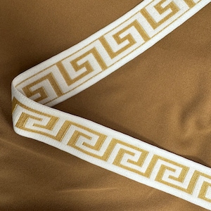 Könnte beinhalten: Weißer elastischer Besatz mit einem goldenen griechischen Schlüsselmuster. Der Besatz befindet sich auf einem braunen Stoffhintergrund.