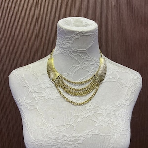 May include: A gold-toned layered necklace with a wide, curved centerpiece and multiple chain strands.