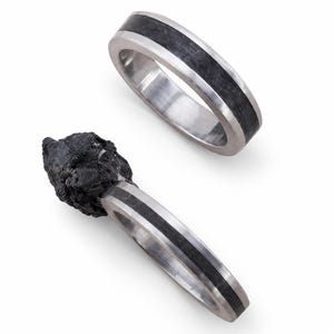 May include: Two rings featuring a polished silver-toned metal band with a central inlay of dark, textured material. One ring is plain, while the other is attached to a jagged, irregular piece of raw black stone that extends from the band.
