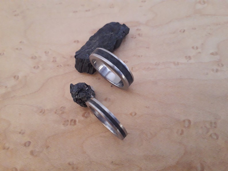 May include: Two silver-toned metal rings featuring a central inlay of dark, textured material. One ring is positioned flat, while the other is tilted upright, resting against a jagged, dark, charcoal-like stone fragment with a rough surface.