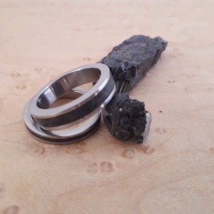 May include: Two silver-toned metal rings featuring a dark, textured inlay band. The rings are positioned next to a jagged, raw piece of black mineral with a crystalline, reflective surface, showcasing a contrast between the smooth metal and stone.