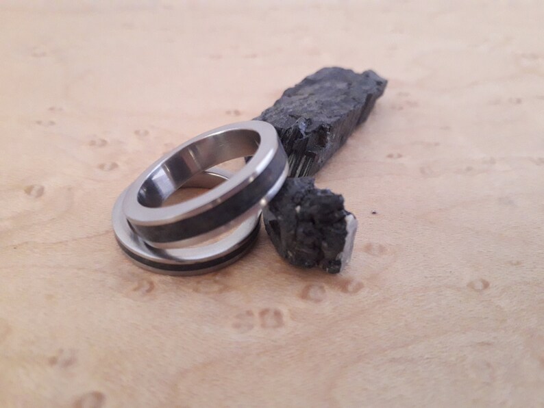 May include: Two silver-toned metal rings featuring a dark, textured inlay band. The rings are positioned next to a jagged, raw piece of black mineral with a crystalline, reflective surface, showcasing a contrast between the smooth metal and stone.