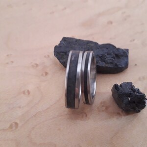 May include: Two silver-toned metal rings featuring a central inlay of dark, textured black material. The rings are positioned upright, accompanied by two jagged pieces of black coal-like rock with a rough, uneven surface texture.