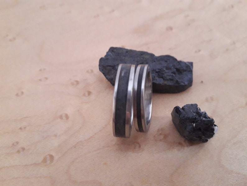 May include: Two silver-toned metal rings featuring a central inlay of dark, textured black material. The rings are positioned upright, accompanied by two jagged pieces of black coal-like rock with a rough, uneven surface texture.
