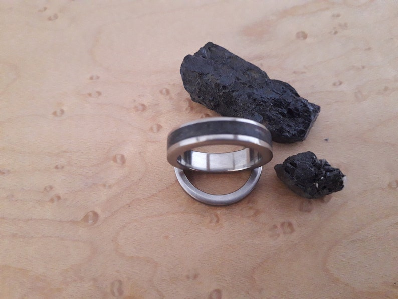 May include: A silver-toned metal ring featuring a dark, textured inlay that runs along the center of the band. The ring is positioned near two jagged, black, coal-like stones, showcasing a smooth metallic interior and a contrasting dark exterior.