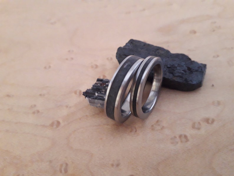 May include: Two silver rings with black inlays. The rings are stacked on top of each other. The rings are on a light wood surface.
