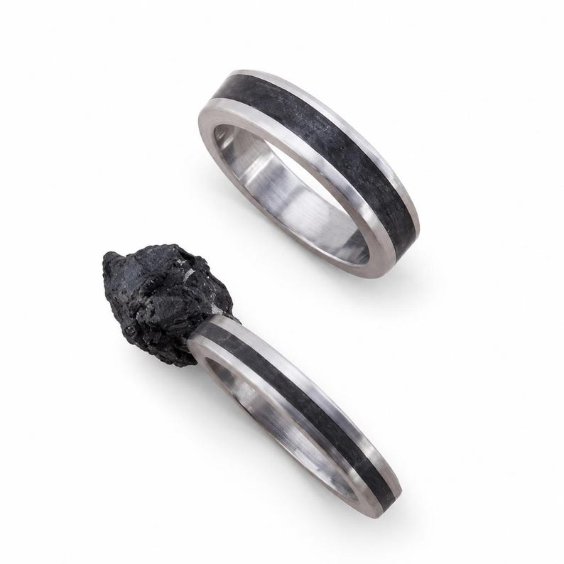 May include: Two rings featuring a polished silver-toned metal band with a central inlay of dark, textured material. One ring is plain, while the other is attached to a jagged, irregular piece of raw black stone that extends from the band.