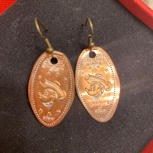 May include: A pair of copper-coloured earrings shaped like a penny with a Mickey Mouse character wearing a pirate hat on one side and the words "Adventureland" on the other side. The earrings have a gold-coloured hook.